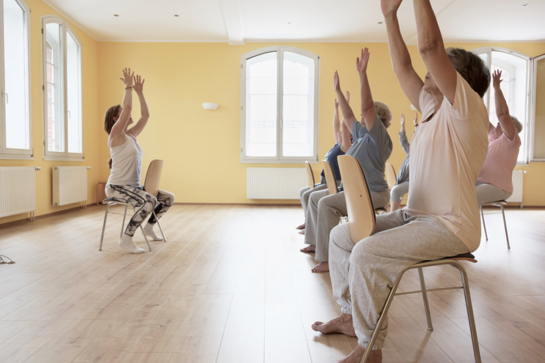 Teacher and active senior women yoga class on chairs, arms raised,